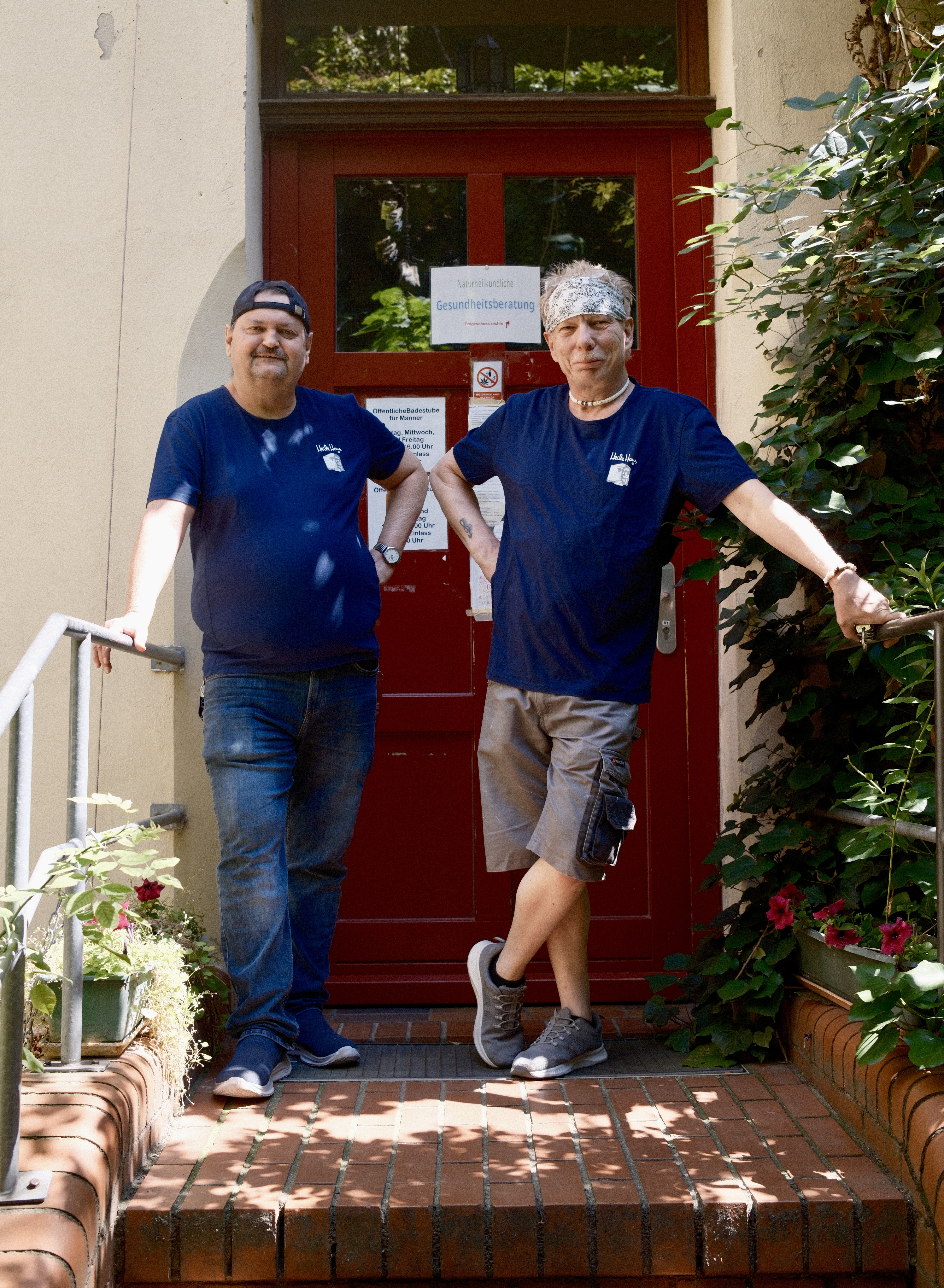 Zwei Mitarbeiter stehen lächelnd draußen vor dem Haus vor dem Duschraum, beide haben ein blaues T-Shirt mit dem Heilehaus Logo an.