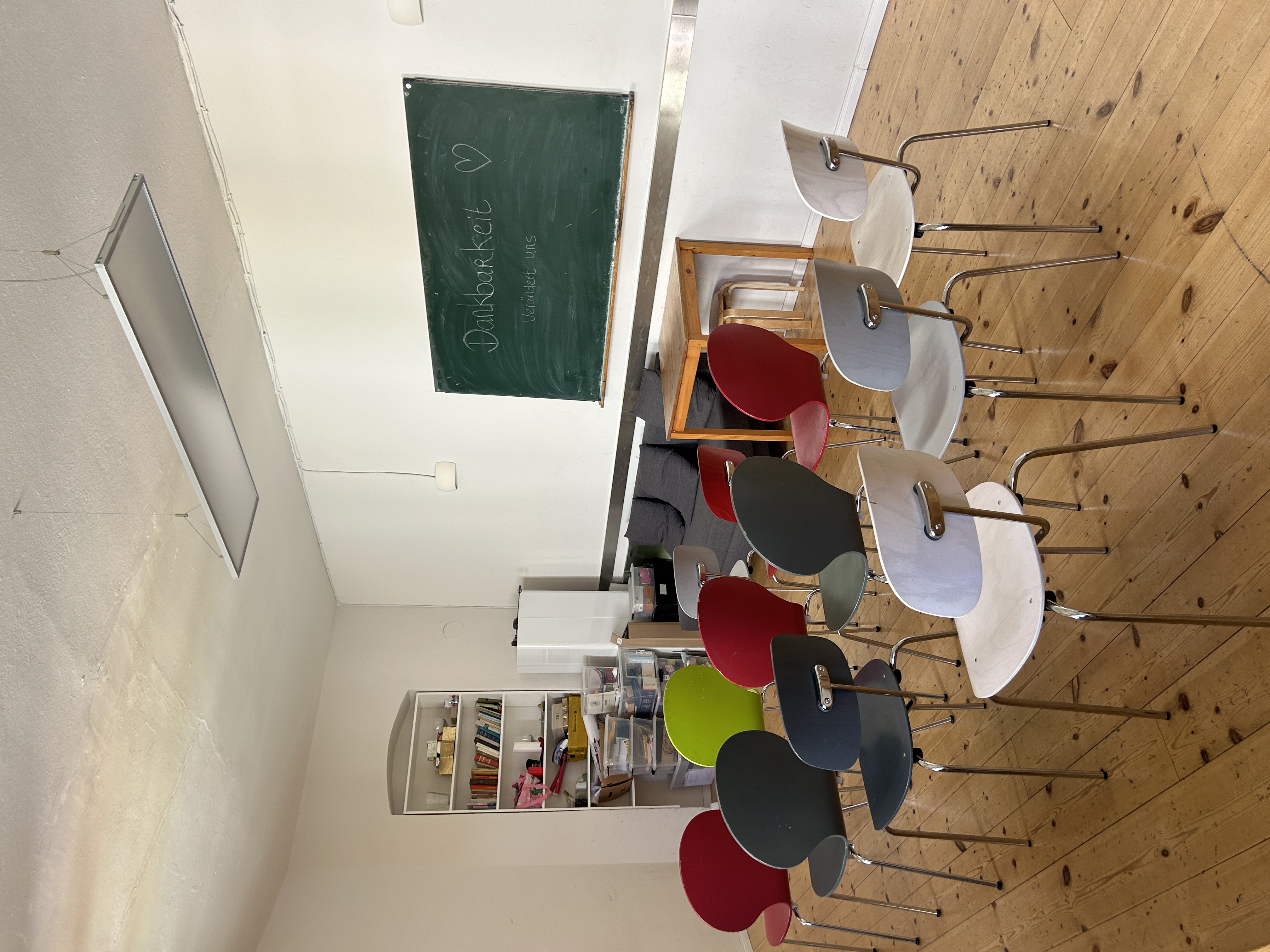 Ein heller und großer Raum mit bunten aufgereihten Stühlen. An der rechten Wand hängt eine grüne Tafel auf der Dankbarkeit mit Kreide geschrieben steht