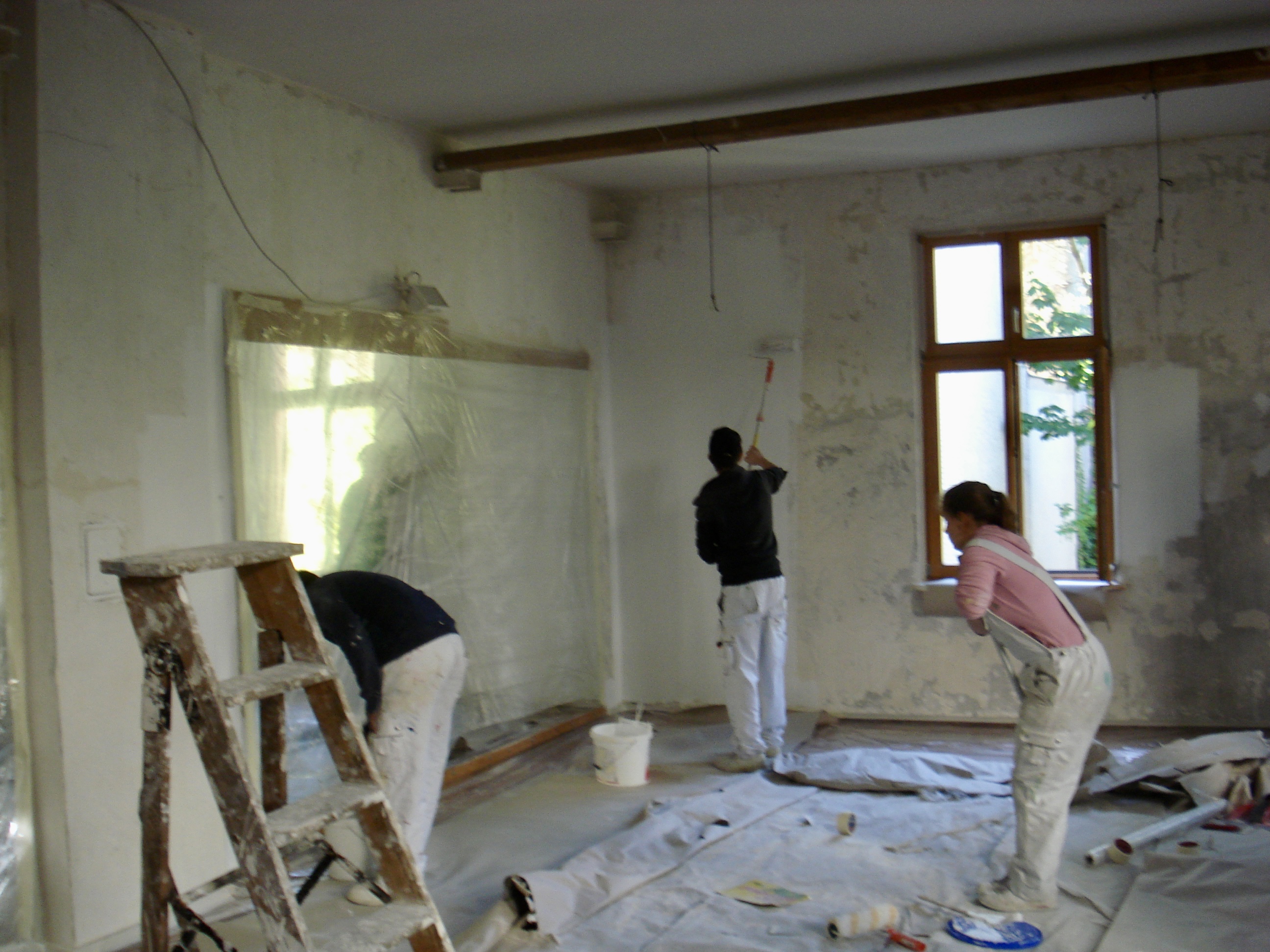 Menschen renovieren einen Raum. Eine Person streicht die Wand, eine andere steht auf einer Leiter. Abdeckfolien und Werkzeug liegen am Boden.