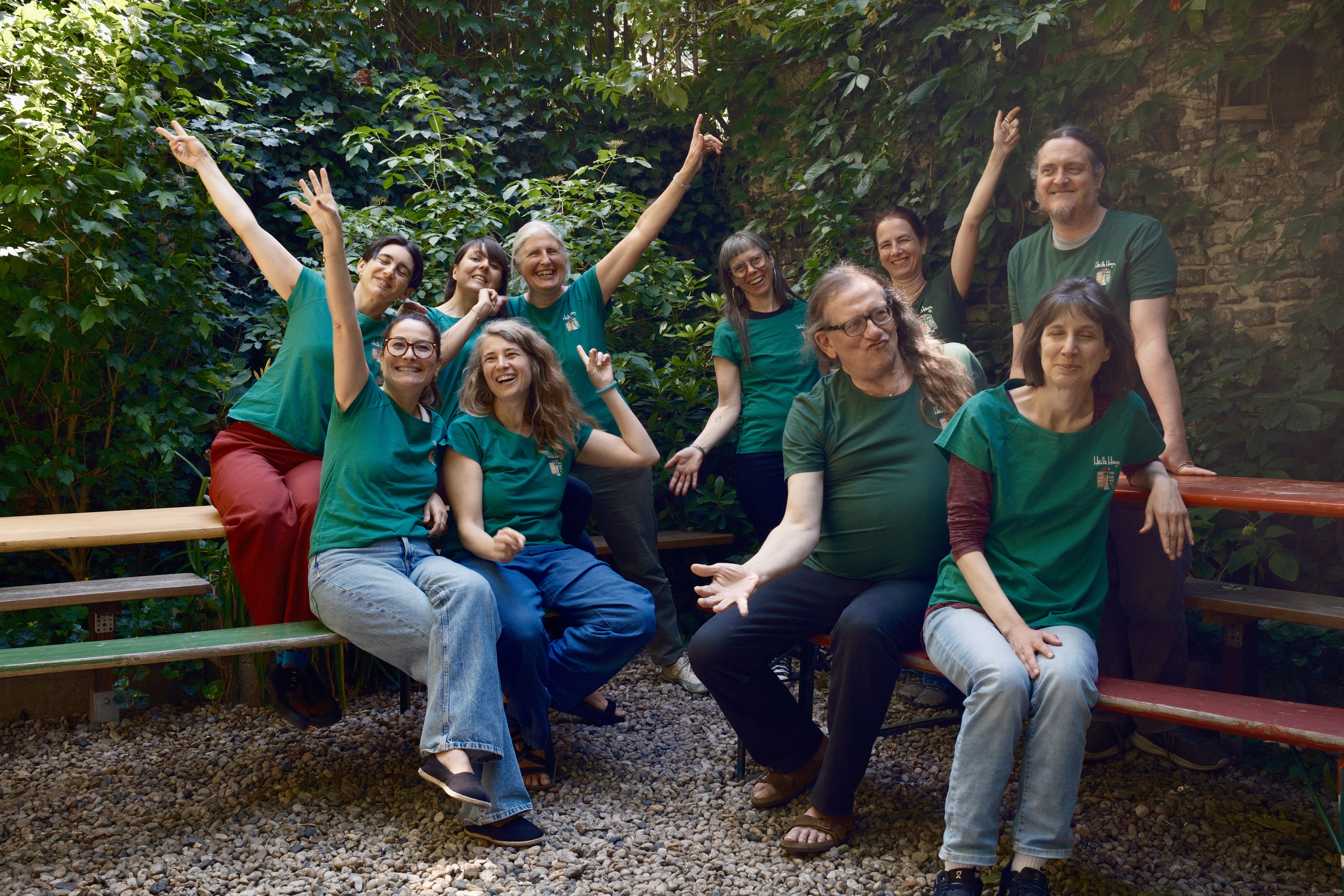 Eine Gruppe von Menschen in grünen T-Shirts mit einem HeileHaus-Logo sitzen und stehen neben einem Tisch fröhlich im Hof. Alle lächeln und heben die Arme.