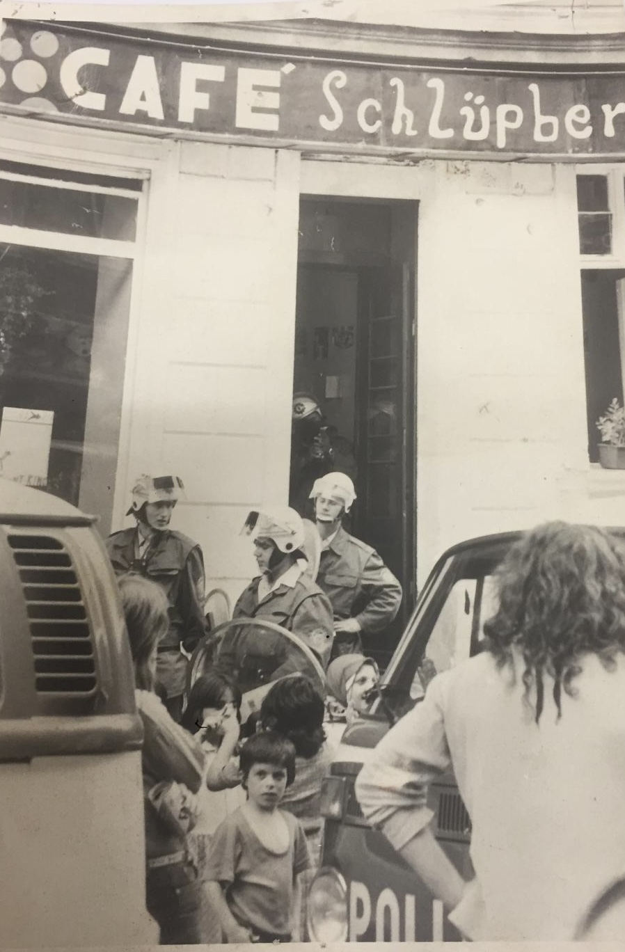 Historisches Schwarz-Weiß-Foto: Kinder und Erwachsene stehen vor einem Gebäude. Mehrere Polizisten sind zu sehen. Das Gebäude zeigt das Logo Café Schlüpber.