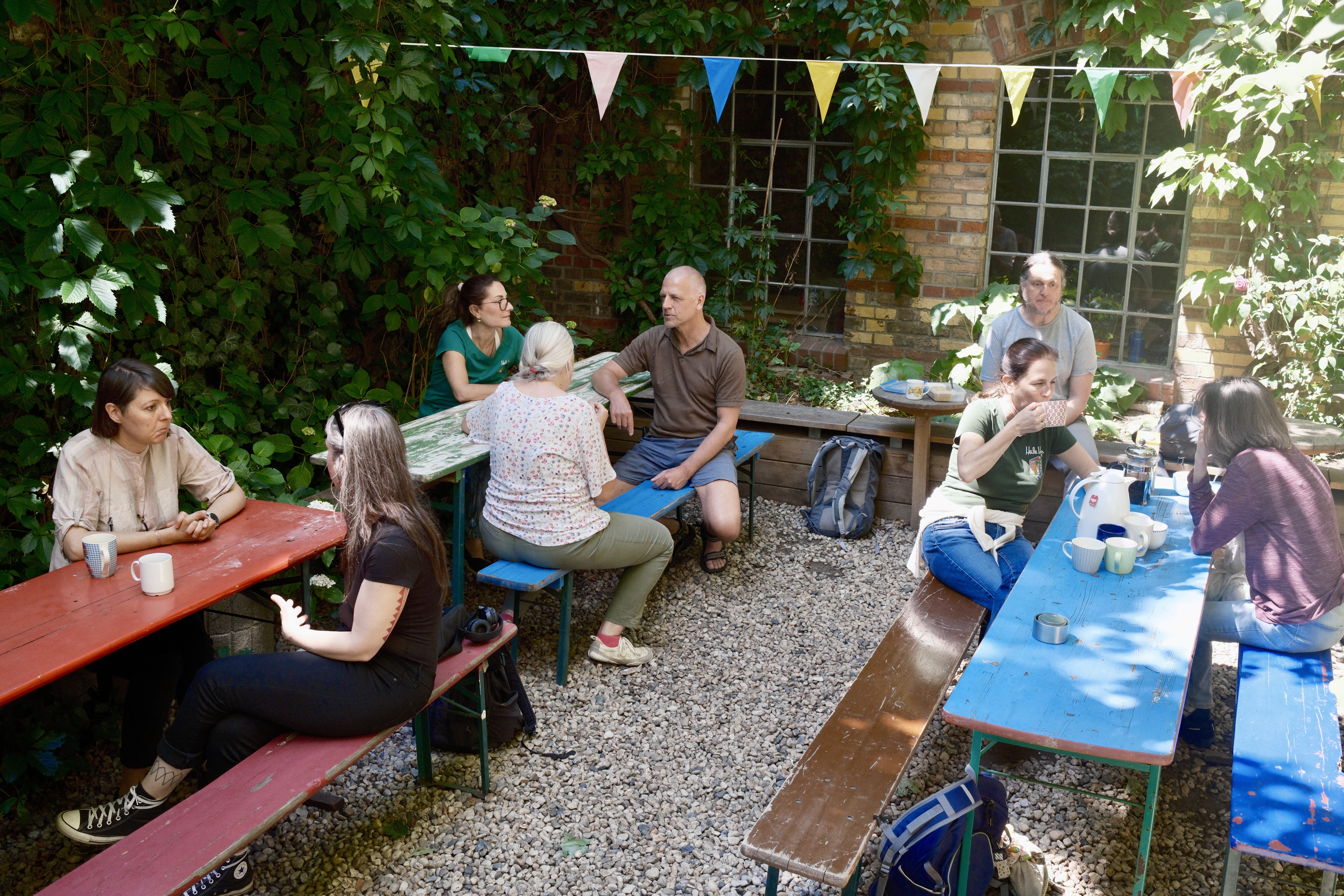 Mehrere Menschen sitzen an bunten Tischen im Hof und sprechen miteinander. Wimpel hängen über dem Sitzbereich.