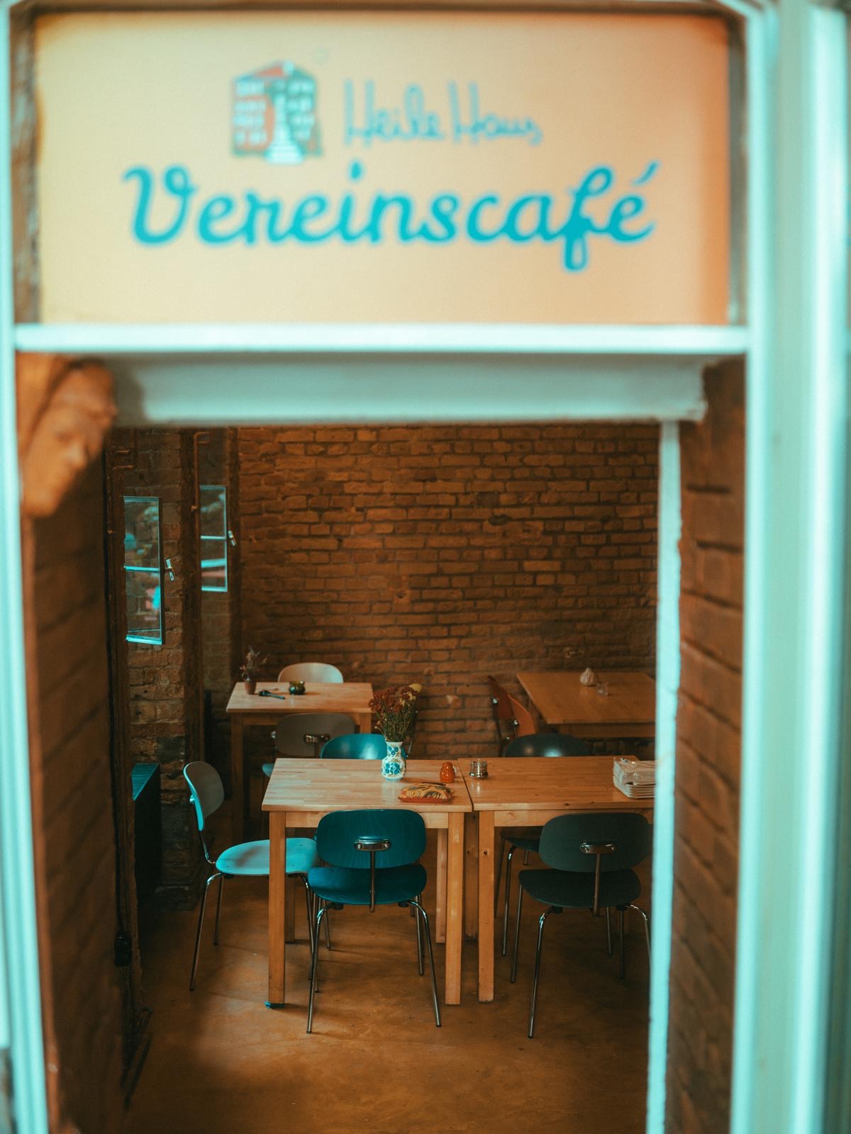 Man sieht den Eingang vom Vereinscafé. Über der Tür hängt das Vereinscafé Logo. Man kann Tische und Stühle erkennen.